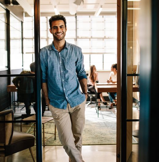 Full length portrait of handsome young man standing in doorway of office with his hands in pockets. Creative male executive at startup with colleagues in background.